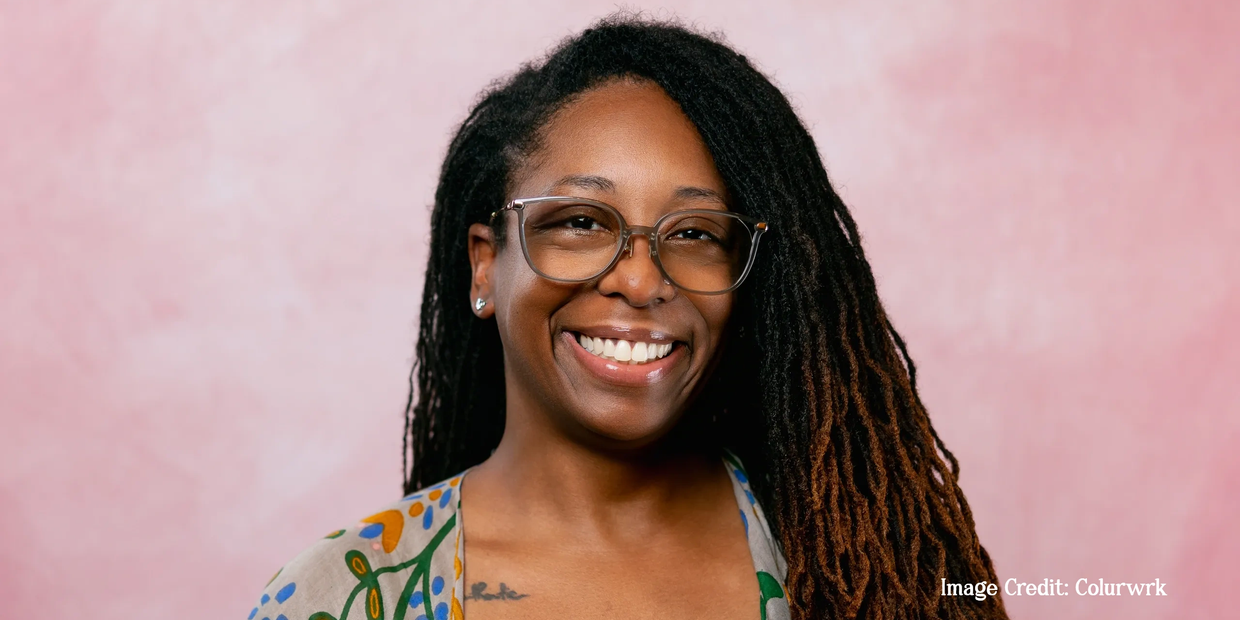 Smiling woman with glasses and long dreadlocks against a pink background.